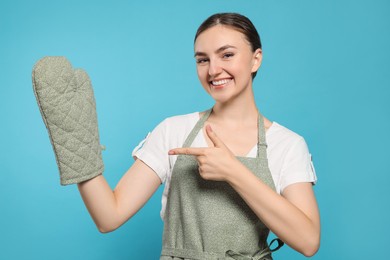 Beautiful young woman in clean apron with pattern and oven glove on light blue background