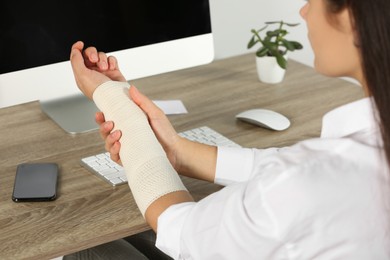 Photo of Young woman with arm wrapped in medical bandage sitting at workplace, closeup
