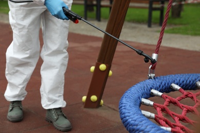 Photo of Woman wearing chemical protective suit with disinfectant sprayer on playground, closeup. Preventive measure during coronavirus pandemic
