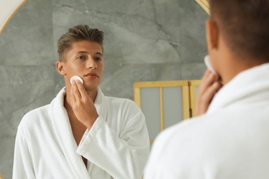 Photo of Handsome young man cleaning face with cotton pad near mirror in bathroom