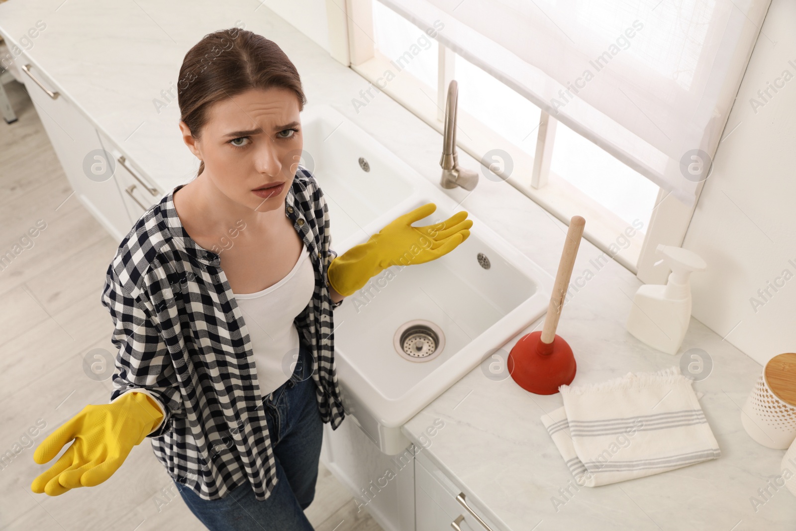 Photo of Upset young woman with plunger near sink in kitchen, above view