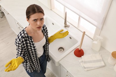 Upset young woman with plunger near sink in kitchen, above view