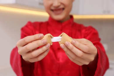 Young woman holding tasty fortune cookie with prediction in kitchen, closeup