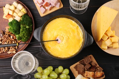 Photo of Fondue pot with melted cheese, fork and different products on black wooden table, flat lay