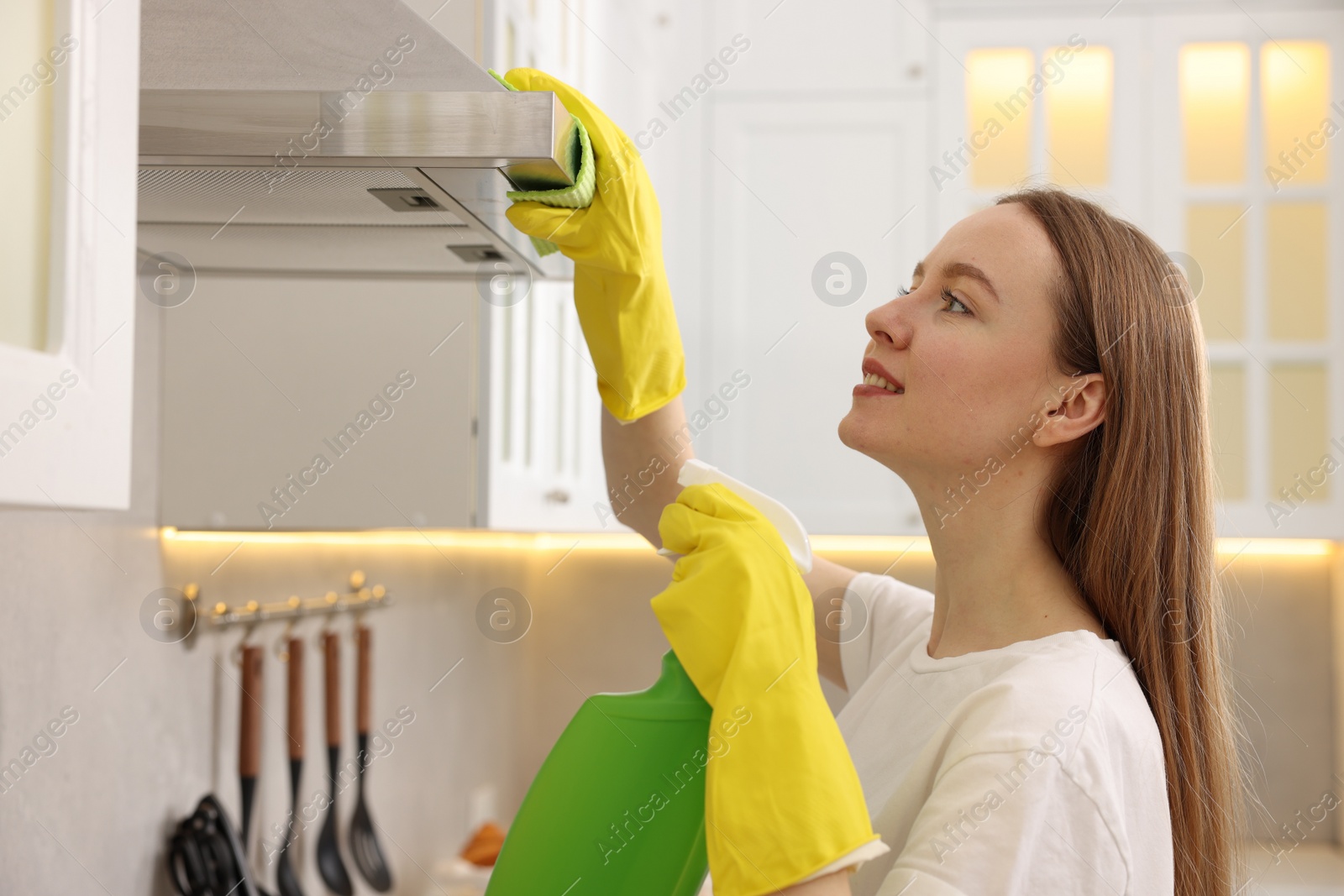 Photo of Woman with spray bottle and microfiber cloth cleaning kitchen hood at home