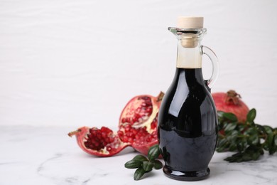 Photo of Tasty pomegranate sauce in bottle, fruits and branches on white marble table, closeup. Space for text