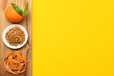 Photo of Wooden board with dried orange zest seasoning, fresh peel and fruit on yellow background, top view. Space for text
