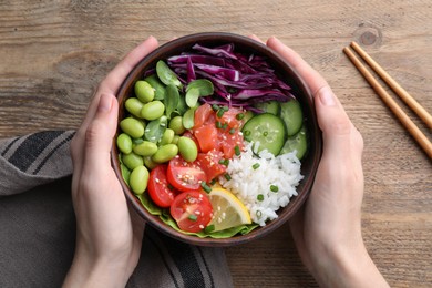 Photo of Woman holding poke bowl with salmon, edamame beans and vegetables at wooden table, top view