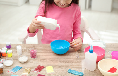 Little girl pouring glue into bowl at table in room, closeup. DIY slime toy