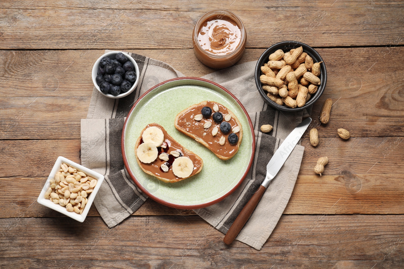 Photo of Toasts with tasty nut butter, banana slices, blueberries and peanuts on wooden table, flat lay