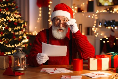Photo of Santa Claus reading letter at his workplace in room with Christmas tree