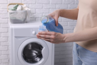 Photo of Woman pouring fabric softener from bottle into cap for washing clothes indoors, closeup