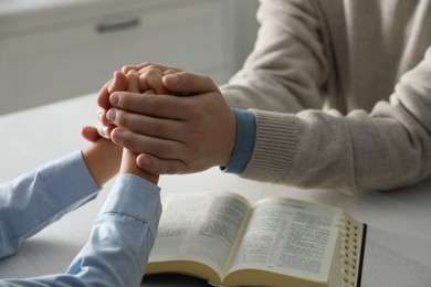 Photo of Boy and his godparent praying together at white wooden table indoors, closeup