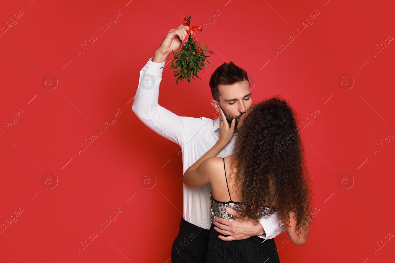 Photo of Happy couple kissing under mistletoe bunch on red background