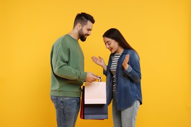 Photo of Man showing shopping bag with purchase to his excited girlfriend on orange background