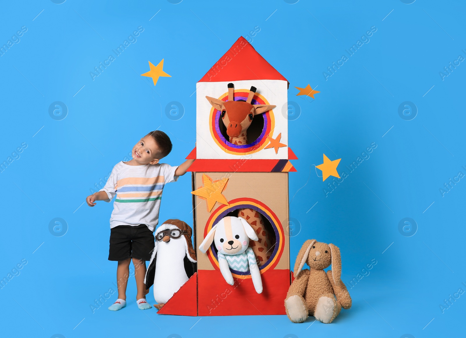 Photo of Cute little boy playing with cardboard rocket and soft toys on light blue background