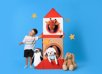 Photo of Cute little boy playing with cardboard rocket and soft toys on light blue background