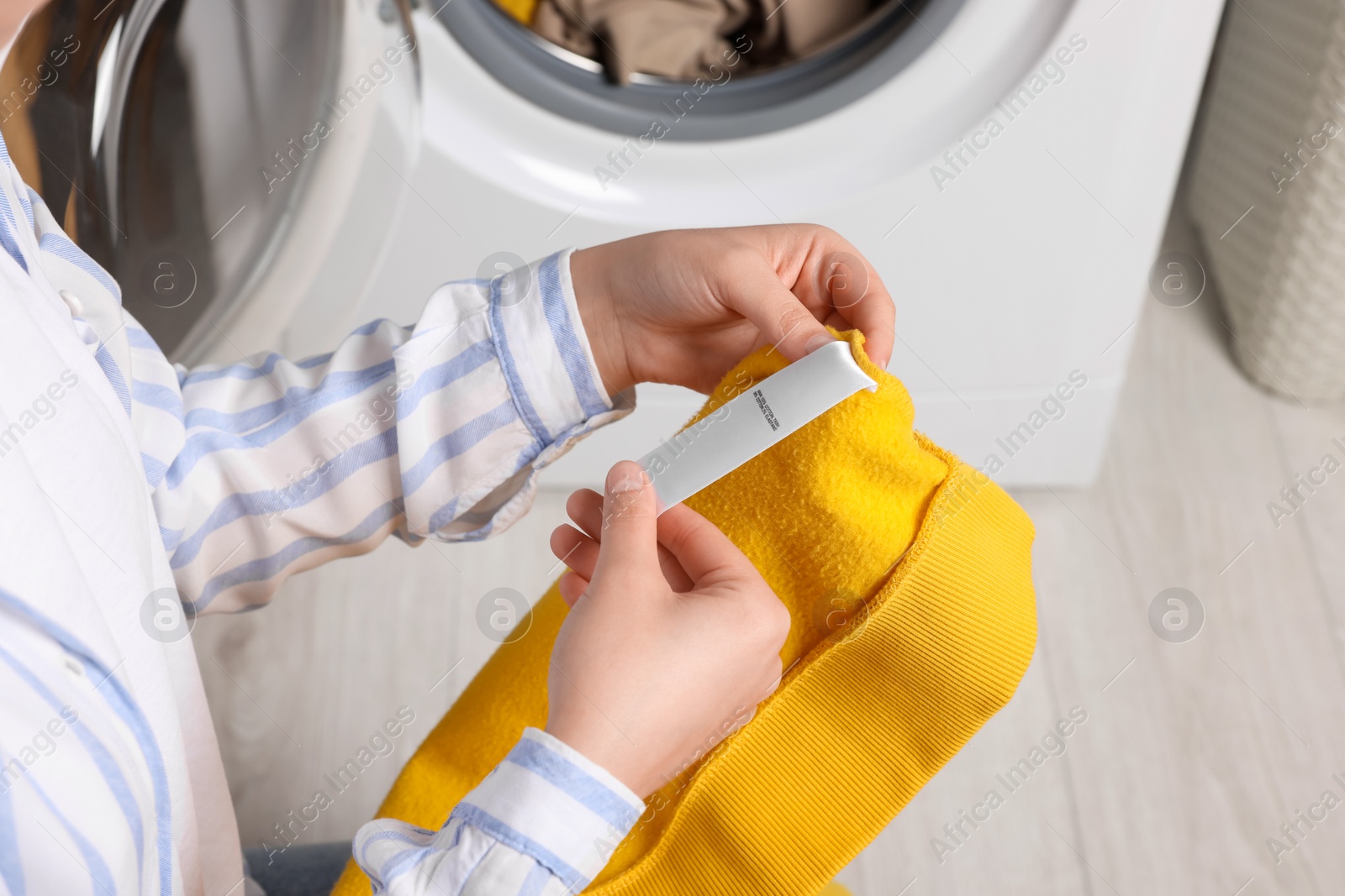 Photo of Woman reading clothing label with care symbols and material content on yellow shirt near washing machine, closeup