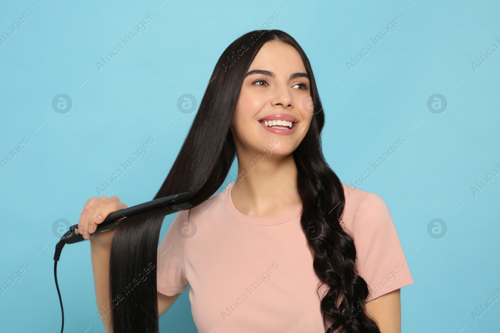 Photo of Beautiful happy woman using hair iron on light blue background