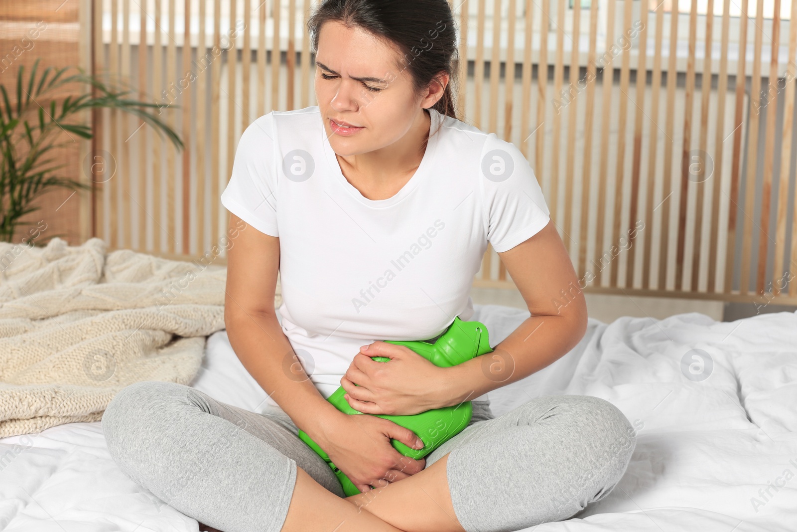 Photo of Young woman using hot water bottle to relieve cystitis pain on bed at home