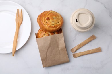 Photo of Paper bag with delicious rolls on white marble table, flat lay. Sweet buns