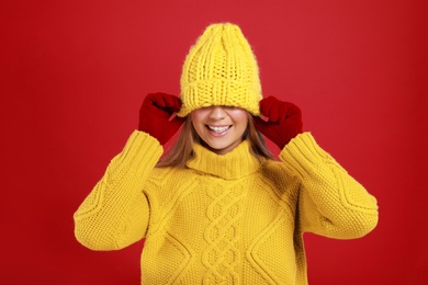 Photo of Funny young woman in warm sweater, gloves and hat on red background. Winter season