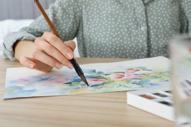 Photo of Woman painting flowers with watercolor at wooden table, closeup. Creative artwork