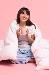 Happy woman with pyjama and blanket holding cup of drink on pink background