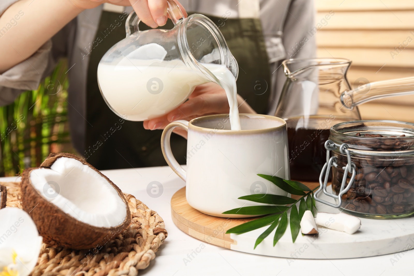 Photo of Woman pouring tasty coconut milk into mug of coffee at white table indoors, closeup