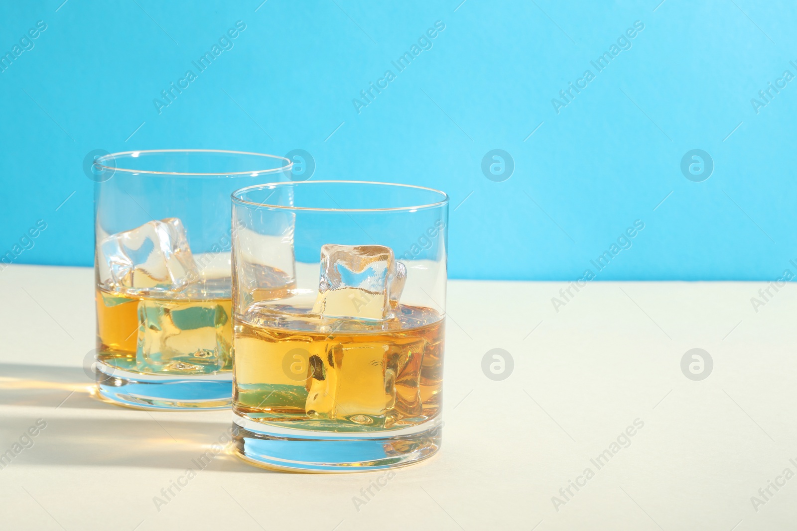 Photo of Whiskey with ice cubes in glasses on white table against light blue background, space for text