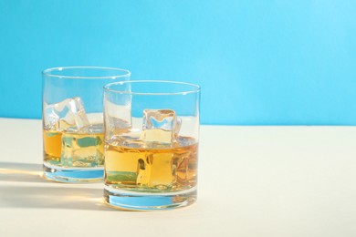 Photo of Whiskey with ice cubes in glasses on white table against light blue background, space for text