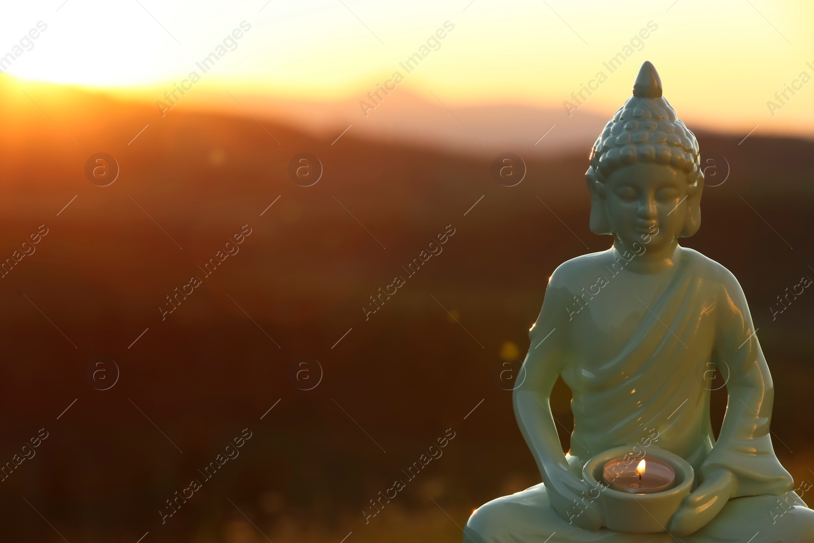 Photo of Decorative Buddha statue with burning candle in mountains at sunset. Space for text