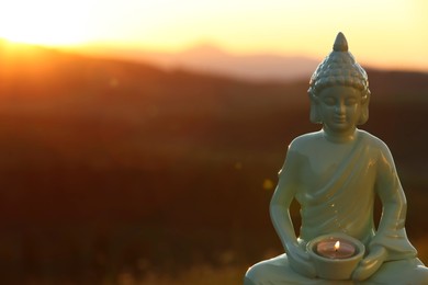 Decorative Buddha statue with burning candle in mountains at sunset. Space for text