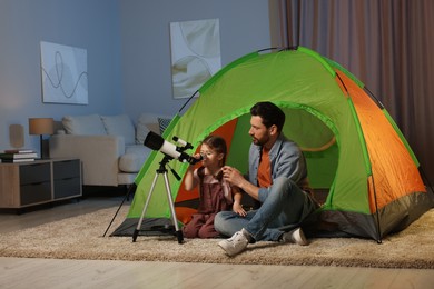 Photo of Father and her daughter looking at stars through telescope while sitting in camping tent indoors