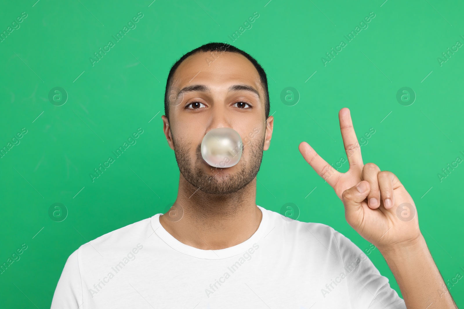 Photo of Portrait of young man blowing bubble gum and showing peace gesture on green background