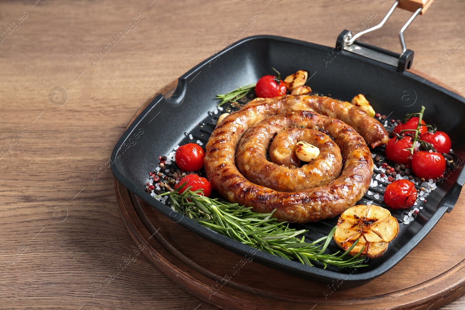 Photo of Delicious homemade sausage with garlic, tomatoes, rosemary and spices in grill pan on wooden table, closeup. Space for text