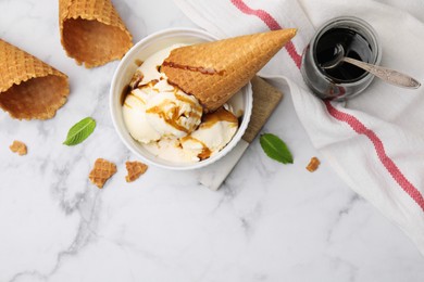 Scoops of ice cream with caramel sauce, mint leaves and cone on white marble table, flat lay. Space for text