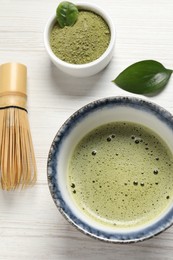 Photo of Cup of fresh matcha tea, bamboo whisk and green powder on white wooden table, flat lay