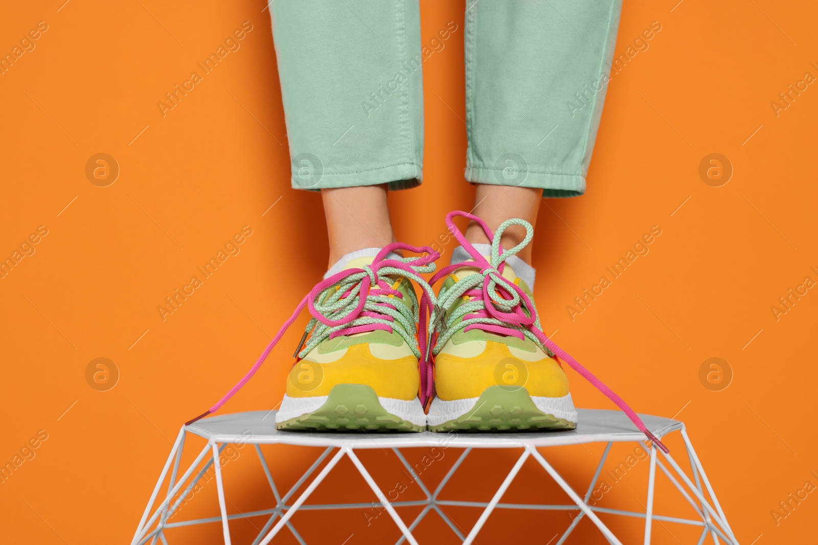 Photo of Woman in stylish colorful sneakers standing on white table against orange background, closeup