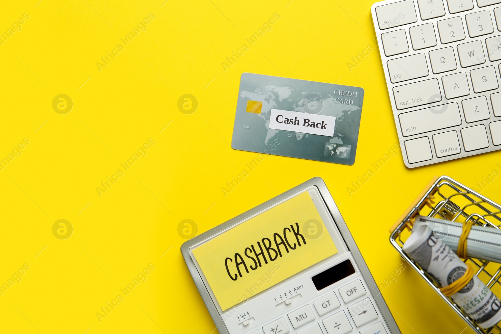 Photo of Calculator, keyboard, credit card and rolled dollar banknotes in shopping cart on yellow background, flat lay with space for text. Cashback concept