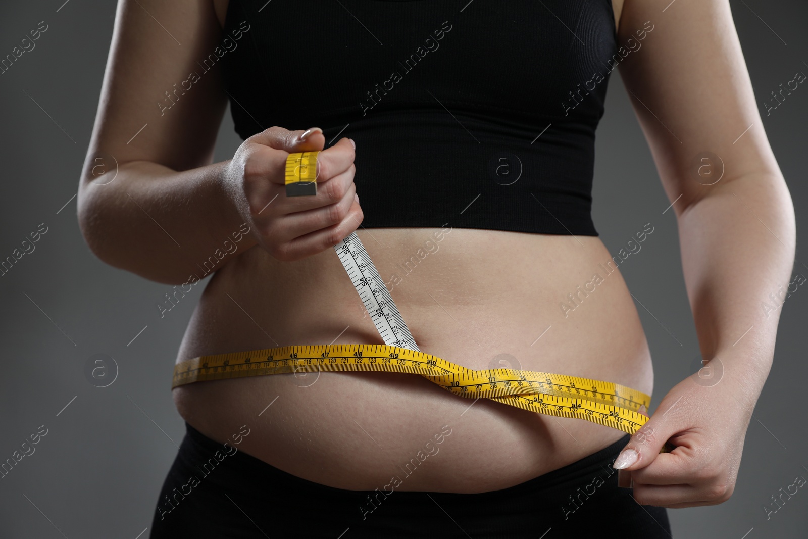 Photo of Woman measuring belly with tape on grey background, closeup. Overweight problem