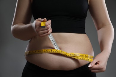 Woman measuring belly with tape on grey background, closeup. Overweight problem
