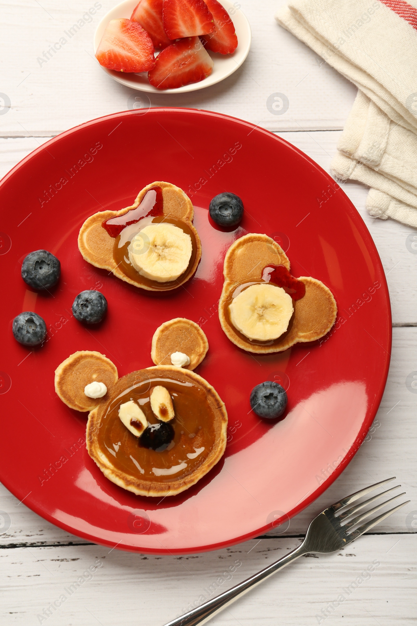 Photo of Creative serving for kids. Plate with cute bears made of pancakes, berries, banana and chocolate paste on white wooden table, flat lay