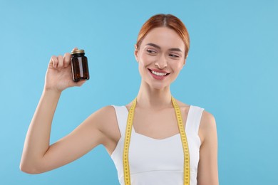 Photo of Happy young woman with bottle of pills and measuring tape on light blue background. Weight loss