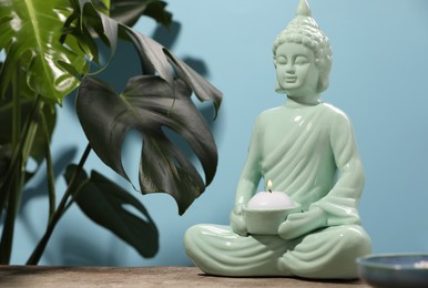 Photo of Buddhism religion. Decorative Buddha statue with burning candle on wooden table and monstera against light blue wall