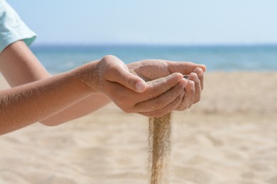 Photo of Child pouring sand from hands on beach near sea, closeup. Fleeting time concept