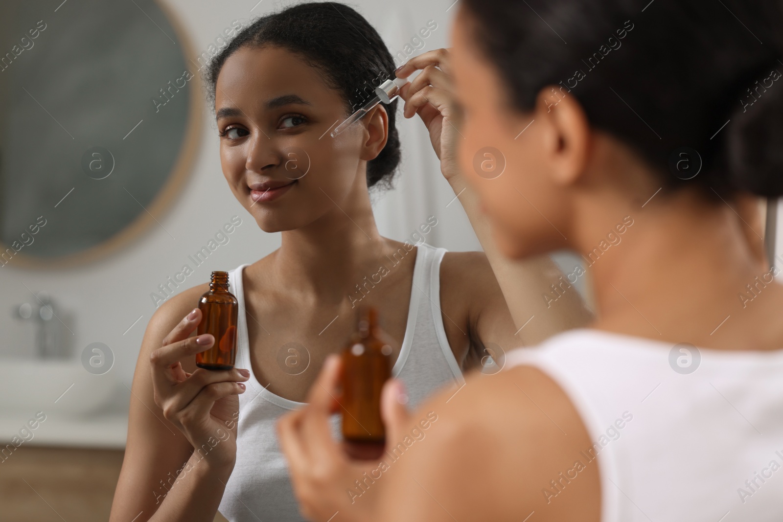 Photo of Beautiful woman applying serum onto her face near mirror in bathroom