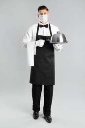 Photo of Waiter in medical face mask holding tray with lid on light grey background