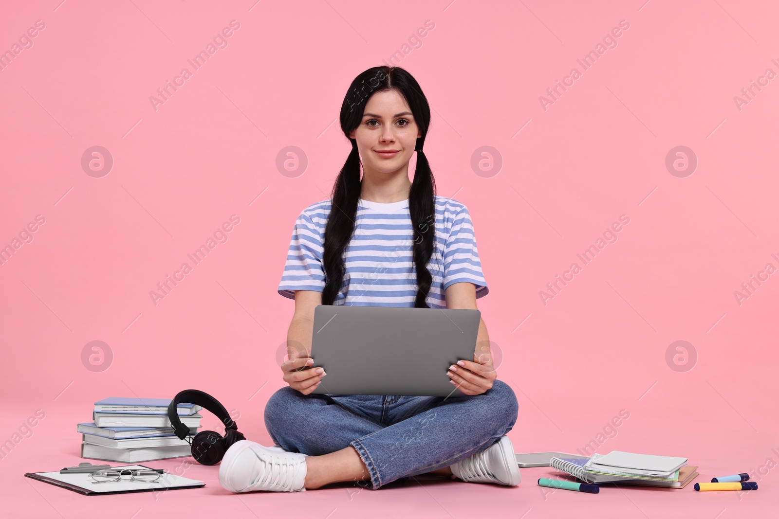 Photo of Student with laptop sitting among books and stationery on pink background