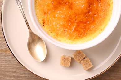 Photo of Delicious creme brulee in bowl, sugar cubes and spoon on wooden table, top view